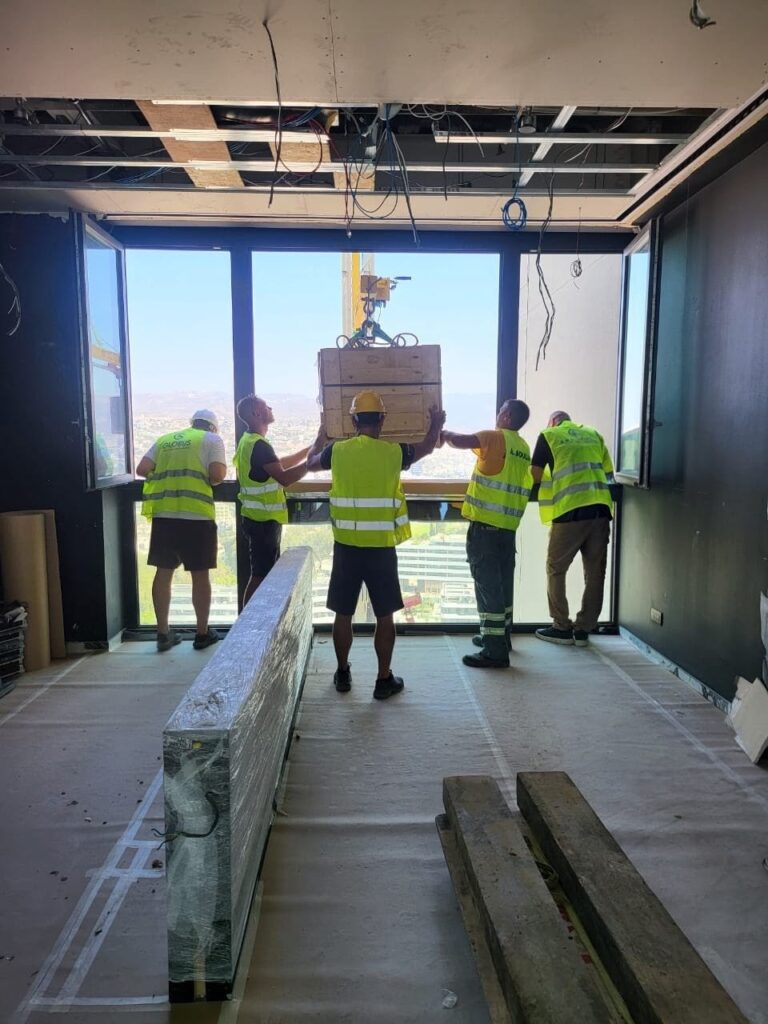 Movers guiding a heavy wooden crate through a high-rise window during a crane-assisted furniture delivery in Limassol.