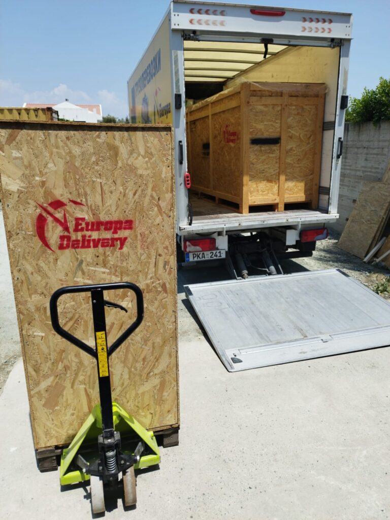 Europa Delivery moving truck equipped with a tail lift and pallet jack, preparing to load a massive wooden crate for international transit.